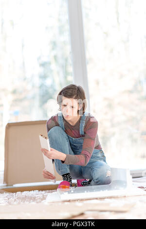 Autorità giovane donna facendo lavori di rinnovo per il fai da te in ginocchio lavorando sul pavimento nel suo salotto di fronte a grandi finestre luminose con copyspace. Foto Stock