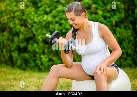 Un sano donna incinta il sollevamento dei pesi sulla palla ginnica in posizione di parcheggio Foto Stock