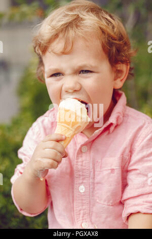 Ritratto di carino caucasico bianco redhead biondo ragazzo di età prescolare con gli occhi blu in maglia rosa mangiare leccare il gelato in cono di cialda Foto Stock