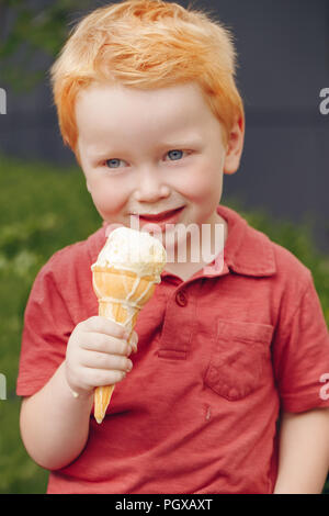 Ritratto di carino caucasico bianco redhead biondo ragazzo di età prescolare con gli occhi blu in t-shirt rossa mangiare leccare il gelato in cono di cialda Foto Stock