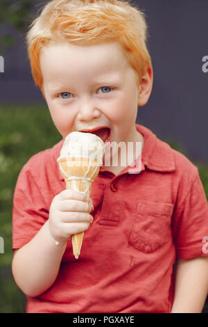 Ritratto di carino caucasico bianco redhead biondo ragazzo di età prescolare con gli occhi blu in t-shirt rossa mangiare leccare il gelato in cono di cialda Foto Stock