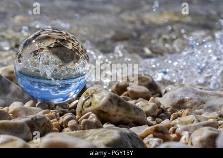 Palla di vetro seduto alla spiaggia rocciosa whit mare onda riflessa in esso/ immagine concettuale delle vacanze estive Foto Stock