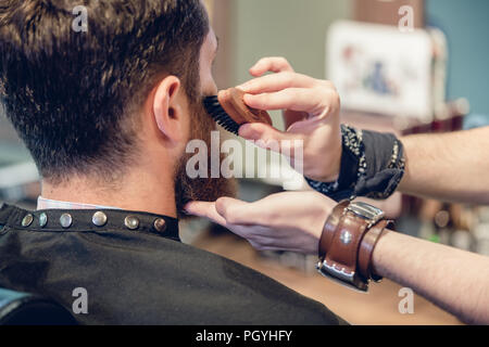 Close-up delle mani di un esperto barbiere usando un handless spazzola con setole di cinghiale, mentre governare la barba del suo giovane cliente in un quartiere alla moda di capelli s Foto Stock