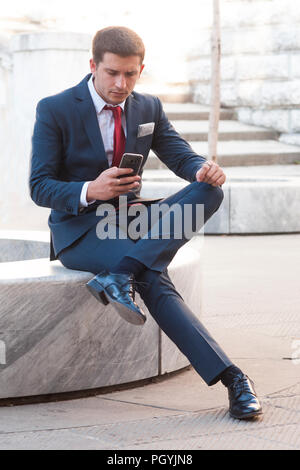 Giovane imprenditore bello in un vestito e un tirante lookink a smartphone seduto su un banco di marmo nel parco Foto Stock