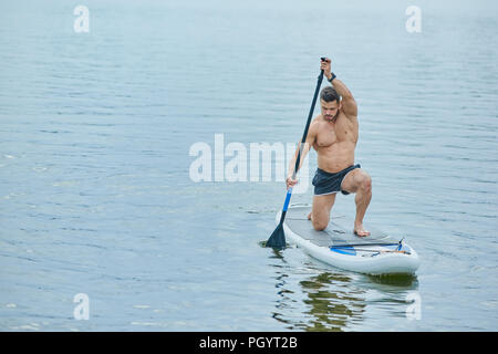 Giovane atleta mantenendo lungo remo in posizione verticale, nuoto sul sup pensione in città sul lago. Modello maschile avente corpo con forti muscoli, mantenendo hea Foto Stock