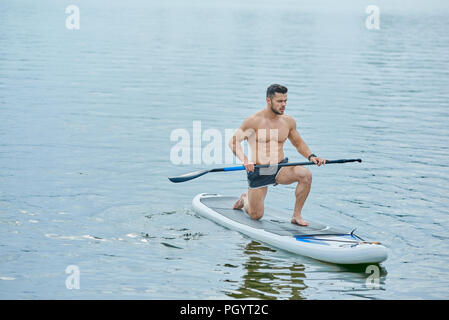 Giovane atleta mantenendo lungo remo in posizione verticale, nuoto sul sup pensione in città sul lago. Modello maschile avente corpo con forti muscoli, mantenendo hea Foto Stock