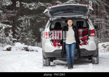 Donna adulta in pelliccia di visone cappotto seduta nel portabagagli della sua auto sotto aperto lo sportello posteriore, foresta invernale Foto Stock