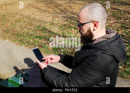 Bel uomo barbuto con gli occhiali è seduta sul banco di verde nel parco e fare affari sul suo smart phone. Foto Stock