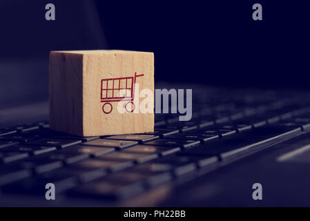 Shopping online e di e-commerce con lo sfondo di un cubo di legno che mostra una icona del carrello appoggiato su di una tastiera di computer visualizzato basso angolo sul nero w Foto Stock