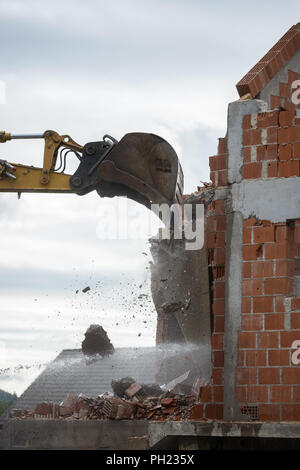 Benna del retroescavatore o escavatore meccanico contro lo skyline demolendo il muro di un edificio in mattoni con battenti in muratura e detriti. Foto Stock