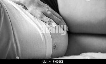 Foto in bianco e nero di donna sdraiata sul letto di toccare la sua gravidanza la pancia e il legame con il suo bambino. Foto Stock