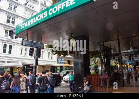 Una grande folla passeggiate passato l'entrata per un caffè Starbucks Cafe di Auckland, in Nuova Zelanda come un uomo seduto di fronte al cafe svolge un tradizionale strumento a corda, 27 febbraio 2018. () Foto Stock