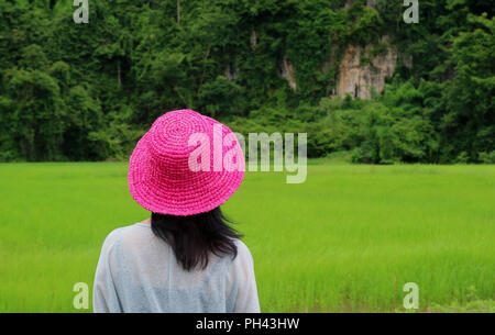 Indossare femmina Vivid Rosa cappello di paglia guardando il verde vivace Campo di riso, durante la stagione delle piogge in phitsanulokb provincia della Thailandia Foto Stock