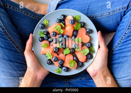 Donna con le mani in mano tenendo la piastra con estate insalata di anguria con mirtilli e foglie di menta sulla lastra grigia. Mangiare sano concetto. Vista superiore Foto Stock