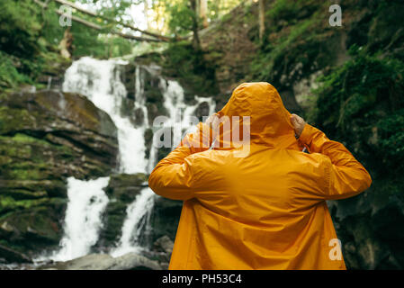 Uomo in piedi in giallo impermeabile e guardando a cascata. escursionismo concept Foto Stock