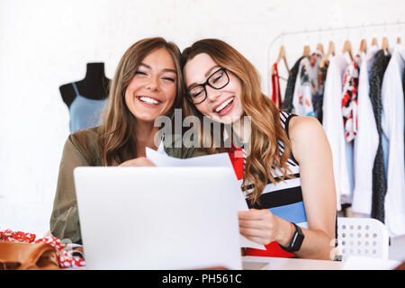 Due allegro di donne giovani stilisti che lavorano insieme presso l'atelier, seduti a tavola con computer portatile e bozze Foto Stock
