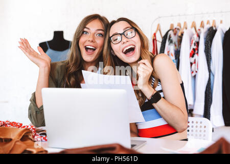 Due lieti di donne giovani stilisti che lavorano insieme presso l'atelier, seduti a tavola con computer portatile e bozze Foto Stock