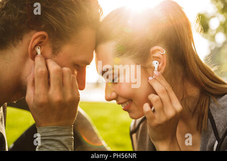 Immagine romantica di coppia giovane uomo e donna 20s in tute ascoltando musica insieme tramite cuffia mentre si passeggia in un parco verde Foto Stock