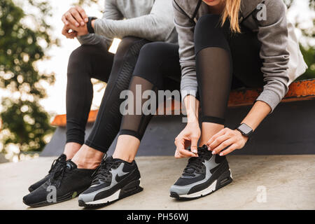 Immagine ritagliata della coppia giovane uomo e donna 20s in tute da seduto sul parapetto in parco verde e legatura lacci delle scarpe Foto Stock