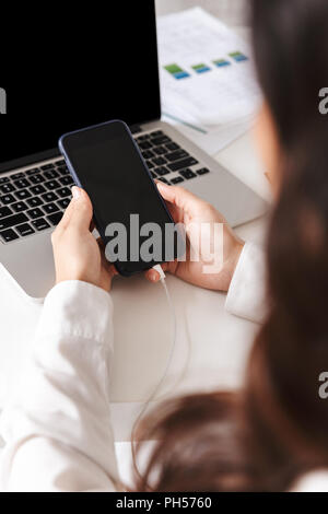 In prossimità di una donna in maglia bianca seduta con il computer portatile su una scrivania, tenendo lo schermo vuoto del telefono mobile Foto Stock