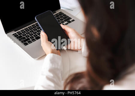 In prossimità di una donna in maglia bianca seduta con il computer portatile su una scrivania in ufficio, tenendo lo schermo vuoto del telefono mobile Foto Stock