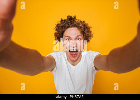 Uomo felice marrone con capelli ricci urlando e prendendo selfie isolate su sfondo giallo Foto Stock