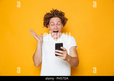 Emozionato uomo europeo in casuale t-shirt bianco urlando mentre guardando smartphone isolate su sfondo giallo Foto Stock