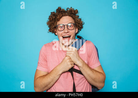 Foto del timido geek ragazzo con capelli ricci con gli occhiali e zainetto sorridente in telecamera isolate su sfondo blu Foto Stock