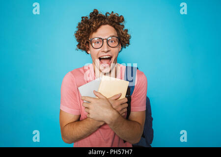 Foto di smart studente ragazzo con capelli ricci con gli occhiali e zainetto sorridente e contenere libri isolate su sfondo blu Foto Stock