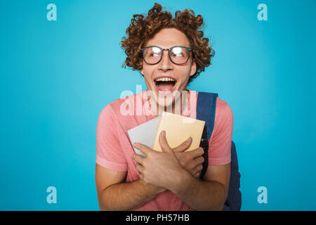 Foto di studente nerd ragazzo con capelli ricci con gli occhiali e uno zaino di ridere e contenere libri isolate su sfondo blu Foto Stock