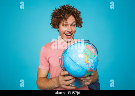 Foto del ragazzo adolescente con capelli ricci indossando uno zaino sorridente e azienda globo terrestre isolate su sfondo blu Foto Stock