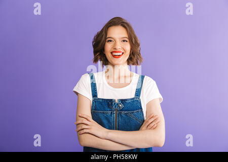 Ritratto di un allegro giovane donna vestito in abiti denim in piedi con le braccia incrociate su sfondo viola Foto Stock