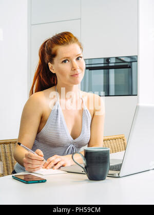 Foto di una bellissima giovane redhead donna seduta in cucina a lavorare sul suo computer portatile e la scrittura di note. Foto Stock