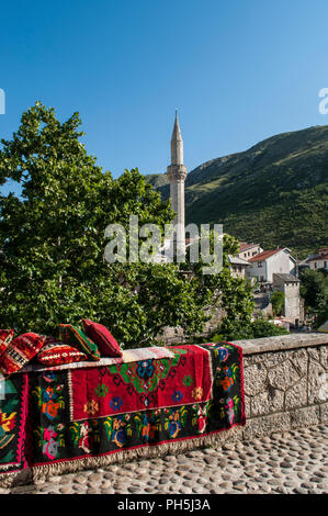 La Bosnia Erzegovina, Europa: lo skyline della città di Mostar con cuscini e tappeti decorati su un muro di pietra e il minareto della Nezir agina moschea dzamija Foto Stock