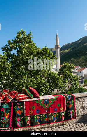 La Bosnia Erzegovina, Europa: lo skyline della città di Mostar con cuscini e tappeti decorati su un muro di pietra e il minareto della Nezir agina moschea dzamija Foto Stock