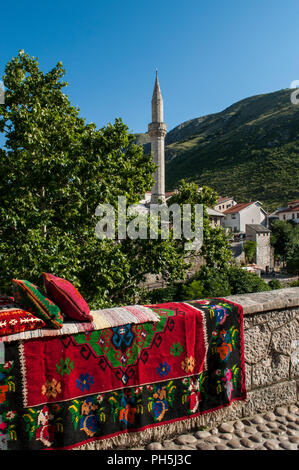 La Bosnia Erzegovina, Europa: lo skyline della città di Mostar con cuscini e tappeti decorati su un muro di pietra e il minareto della Nezir agina moschea dzamija Foto Stock