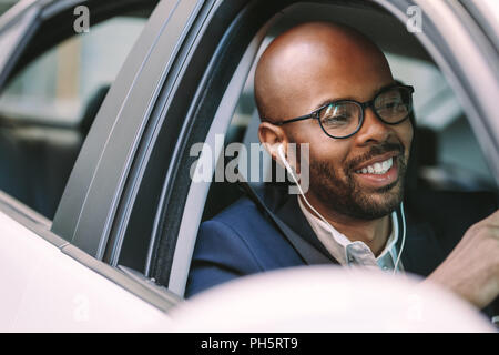 Giovane uomo calvo sorridente dentro la macchina durante la guida. L'uomo africano in tuta e indossare gli auricolari godendo di guida di un'auto. Foto Stock