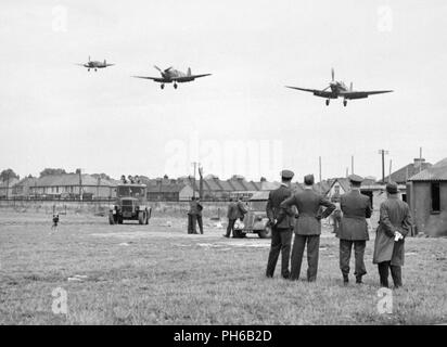 Il personale del No.121 (Aquila) Squadron guarda su come tre Supermarine Spitfire aeromobile atterra dopo un combattente di spaziare su nel nord della Francia a Royal Air Force Rochford in Essex, Inghilterra, Agosto 1942. Alcuni alloggi utilizzati dallo squadrone è visibile in background, come sono diverse case di civili e due veicoli RAF. Foto Stock