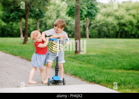 Due piccoli caucasica i bambini in età prescolare combattimenti di colpire ogni altro. Un ragazzo e una ragazza non può condividere uno scooter. I vecchi fratello gemello di non dare il suo giocattolo Foto Stock