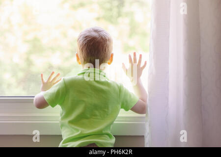 Ritratto di bianco bambino caucasico boy a casa guardando fuori della finestra all'esterno. Vista dal retro. Ragazzo in attesa di qualcuno Foto Stock