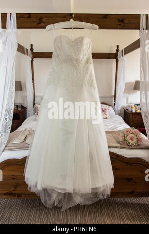 Abito da sposa appeso su un letto a baldacchino in attesa di essere indossato da una sposa il giorno delle nozze, prese alla luce del giorno in una camera da letto Foto Stock