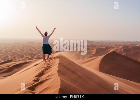 Escursionista femmina solleva le braccia in aria, red dune di sabbia nel deserto, paesaggio di dune Erg Chebbi, Merzouga, Sahara, Marocco Foto Stock