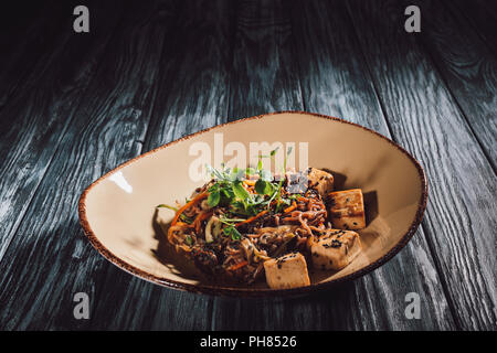 Vista ravvicinata di soba con tofu e verdure decorate con semi germinati di girasole su piastra su un tavolo di legno Foto Stock