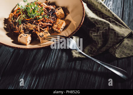 Angolo di alta vista di soba con tofu e verdure decorate con semi germinati di girasole su piastra in prossimità di asciugatutto e forcella su un tavolo di legno Foto Stock