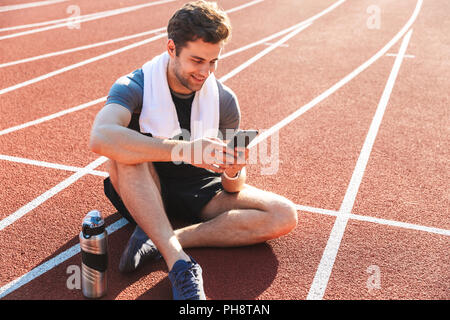 Sorridente sportivo finito l'esecuzione presso lo stadio, riposo, utilizzando il telefono cellulare Foto Stock