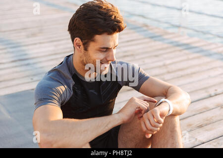 Foto di sorridente uomo sportivo 20s in tuta seduti sul tappeto e guardando il cronometro dopo allenamento sul molo in legno al mare in mattinata Foto Stock