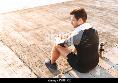 Foto di adulto sportivo 20s in tuta l'ascolto di musica attraverso le cuffie senza fili mentre è seduto sul molo in legno con il thermos tazza in mare Foto Stock