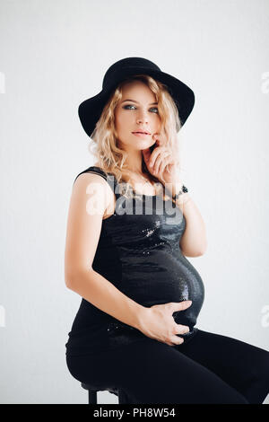 Elegante donna incinta in black hat e frizzante top abbracciando il suo ventre. Foto Stock