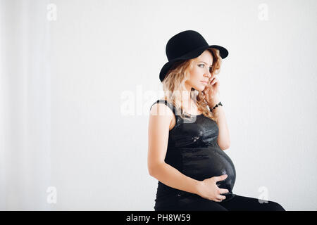 Elegante donna incinta in black hat e frizzante top abbracciando il suo ventre. Foto Stock