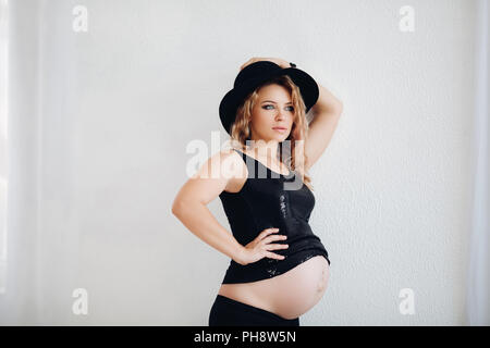 Elegante donna incinta in black hat e frizzante top abbracciando il suo ventre. Foto Stock